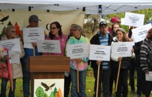 A group of tenants stand under a tent holding signs stating "Our homes | Our community" and "not one more tree." There's a podium in front of them with the Riveria Estates Tenant Union Logo on the front featuring a cluster of trees with an eagle flying over it.