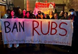 Photo of tenants in a brick building grinning and holding up a white banner with the words "BAN RUBS" written on it. In the background there is another red banner for Puget Sound Tenant Union.
