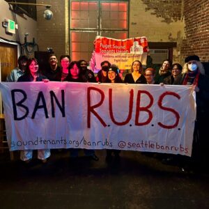 Photo of tenants in a brick building grinning and holding up a white banner with the words "BAN RUBS" written on it. In the background there is another red banner for Puget Sound Tenant Union.