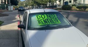 Image of a white vehicle with a green sign in the windshield reading "DO NOT RENT HERE" parked in front of a complex with construction. 